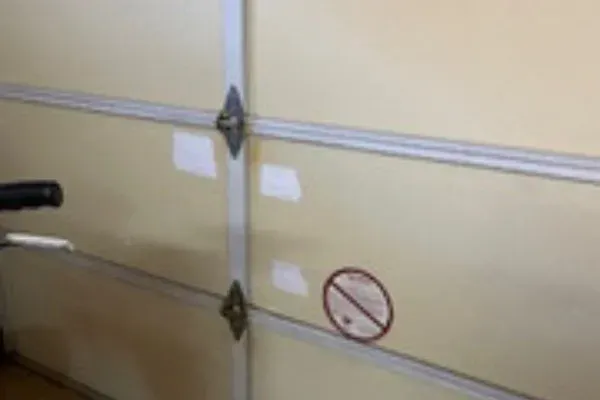 A close-up view of a beige garage door section with horizontal metal panels and a circular red warning sticker.