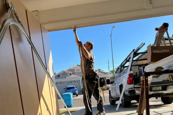 A male technician works on garage door rails, viewed from a low angle, with a work truck and house in the background.